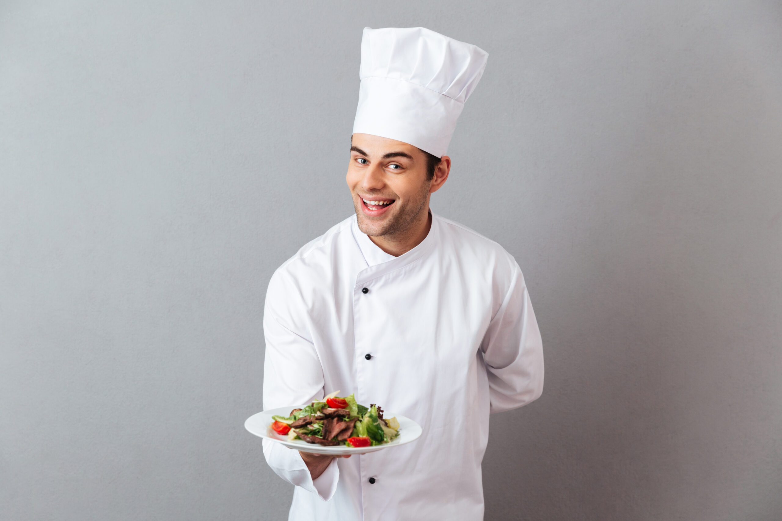 Picture of happy young cook in uniform standing isolated over grey wall background. Looking camera holding salad.